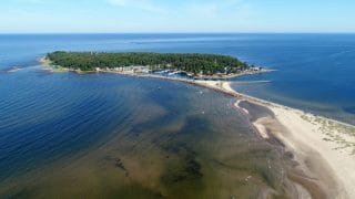 Lintuperspektiivistä otettu kuva meren ympäröimästä Ohtakarista ja sen venesatamasta. Etualalla näkyy pieni palanen hiekkarantaa sekä Ohtakariin vievä kapea tie. Ohtakarin venesataman takana kasvaa metsää.
